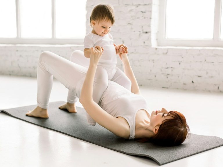 Eine Mutter macht Yoga Übungen auf der Matte, während sie ihr Kind spielerisch anhebt.